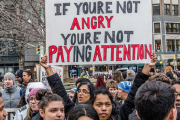 A large gathering of multi-racial young adults at a protest on the streets of a city. In the centre a woman holds a large sign on which the words "If you are not angry you are not paying attention" have been painted