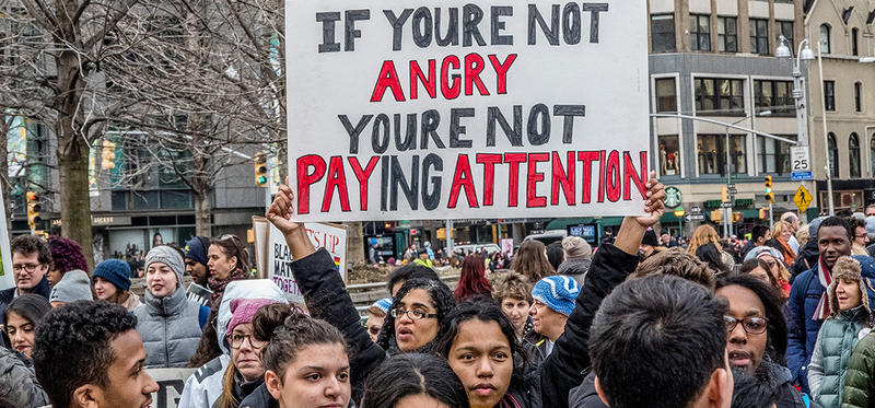 A large gathering of multi-racial young adults at a protest on the streets of a city. In the centre a woman holds a large sign on which the words "If you are not angry you are not paying attention" have been painted