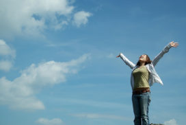 A young woman stands on a cliff top with her arms wide and looks towards a blue sky with wispy clouds