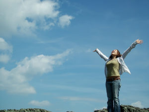 A young woman stands on a cliff top with her arms wide and looks towards a blue sky with wispy clouds